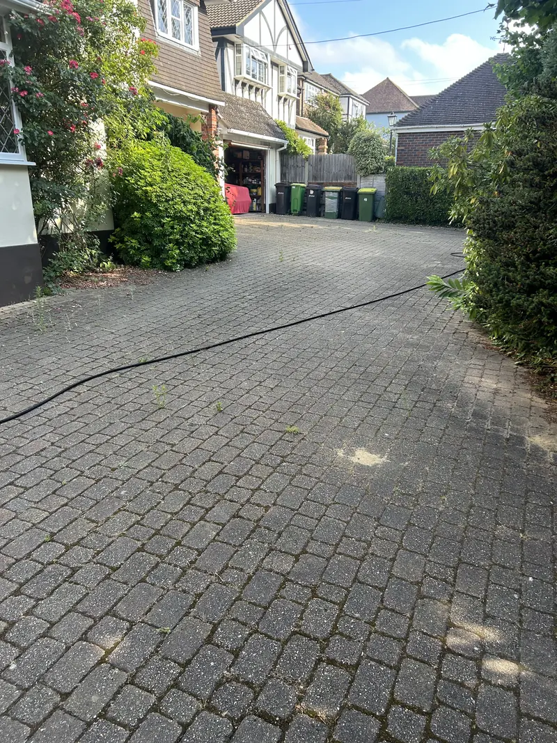 Long grey block-paved driveway during pressure washing, hose across the paving